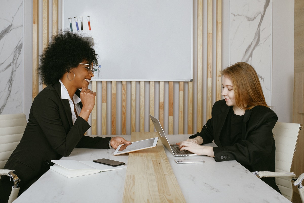 Two women sitting at a desk working on a laptop and tablet with a white board behind them.