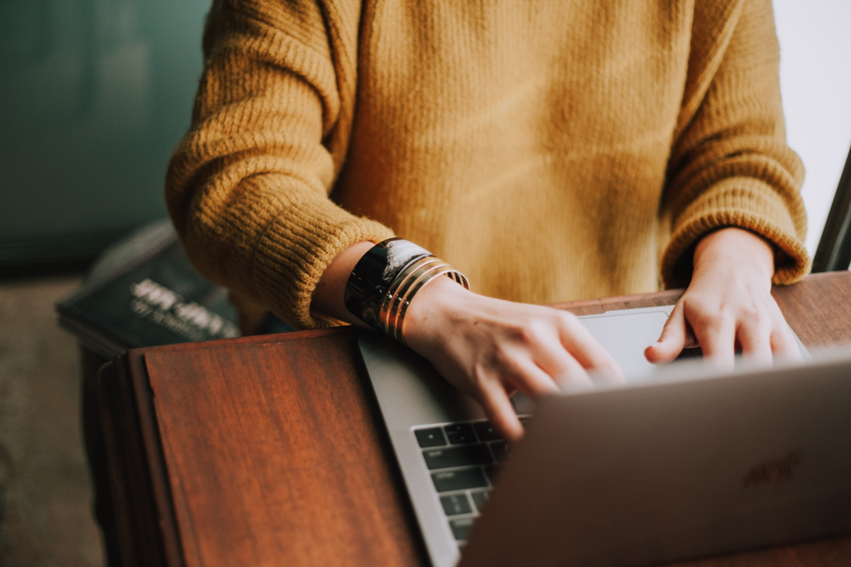 Woman in a yellow sweater with bracelets on her arms working on a silver computer that is sitting on a wood table.