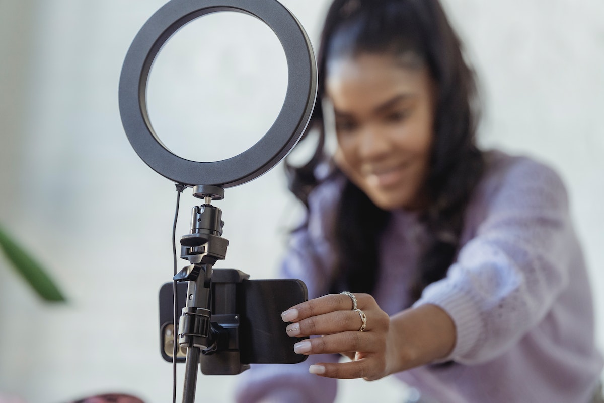 A black woman in a purple sweatshirt filming an ad, sponsored post with a phone set up on a ring light stand.