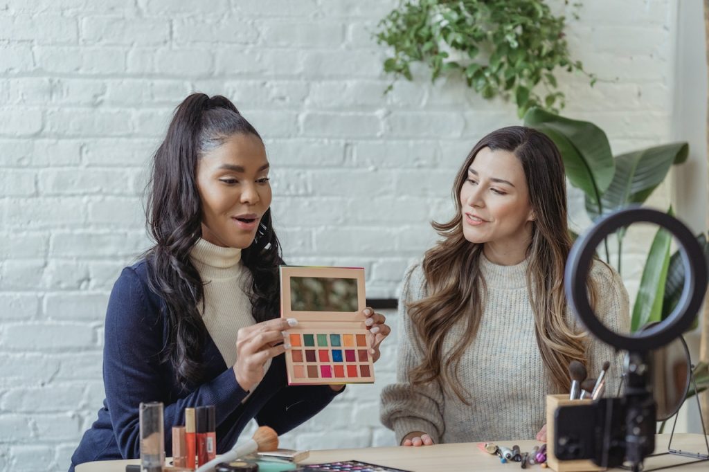 Influencers sitting at a table in front of a camera and ring light showing makeup products.