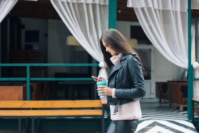 Woman walking by a store holding a coffee cup nad looking at the BeReal app on her phone.