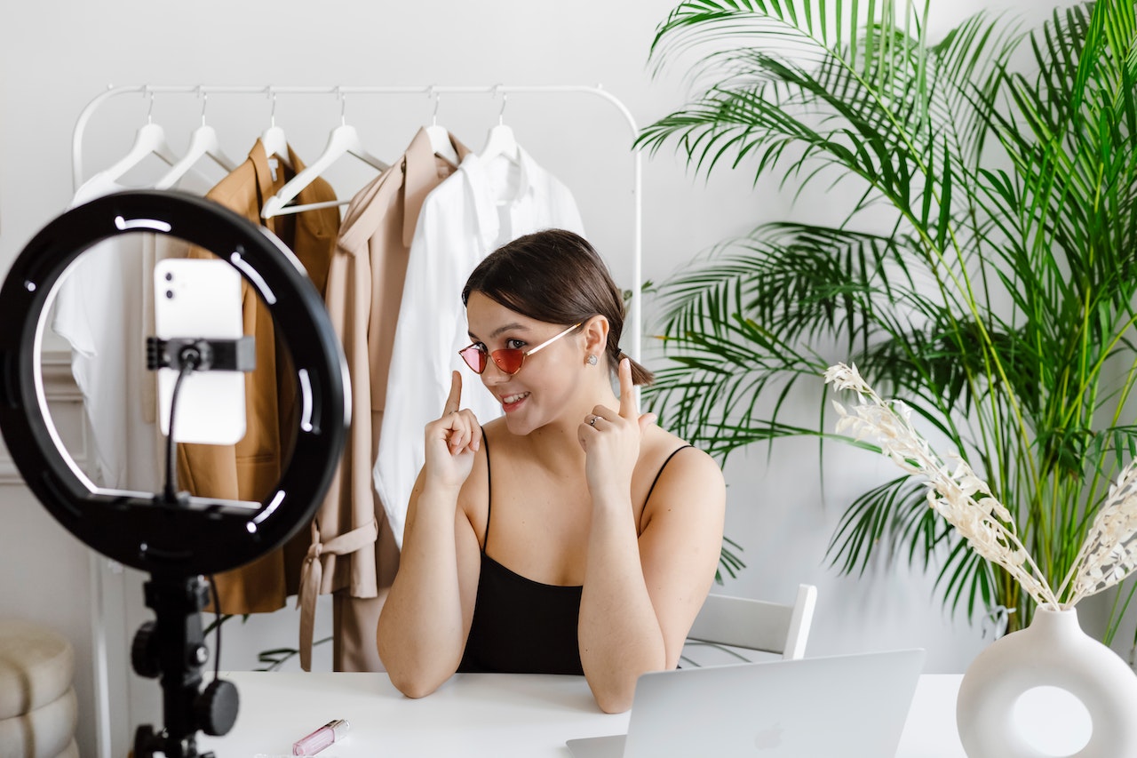 Woman sitting at a desk wearing sunglasses with a ring light and phone in front of her filming. 