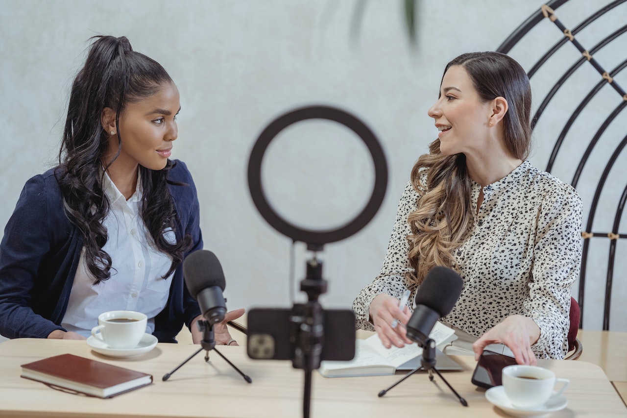 A black woman and a while woman sitting at a desk with microphones and a ring light in front of them recording influencer content.