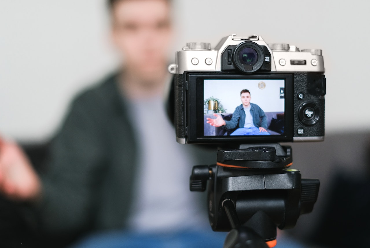 A camera set up on a tripod recording a man sitting on a couch behind it. 