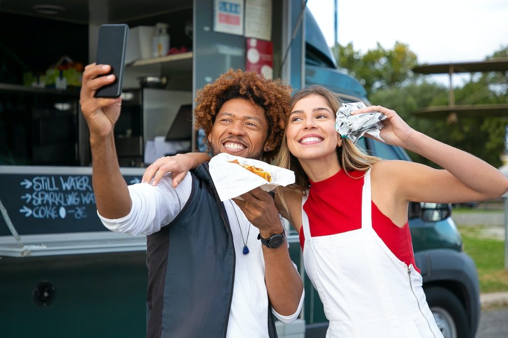 Man and woman standing in front of a food truck taking a picture with their food.