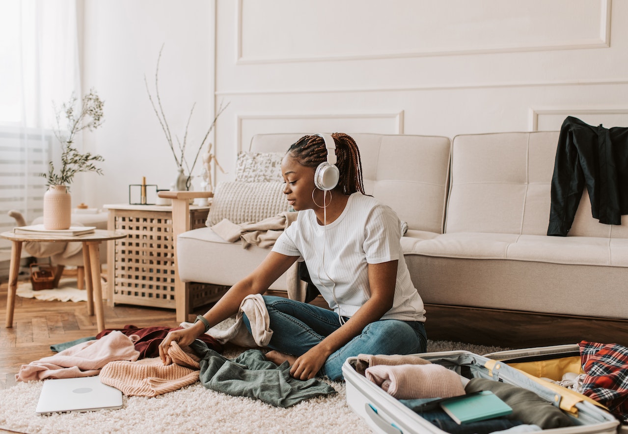 A black woman who is a travel influencer with headphones on her ears sitting on the floor in front of a couch packing a suitcase.