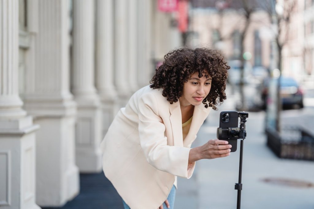 Woman wearing a white jacket adjusting a phone on a tripod to make a YouTube Shorts video on the city street.