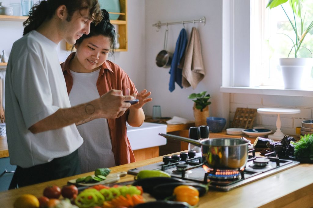 Man and woman in the kitchen cooking and crafting social media posts on a phone.