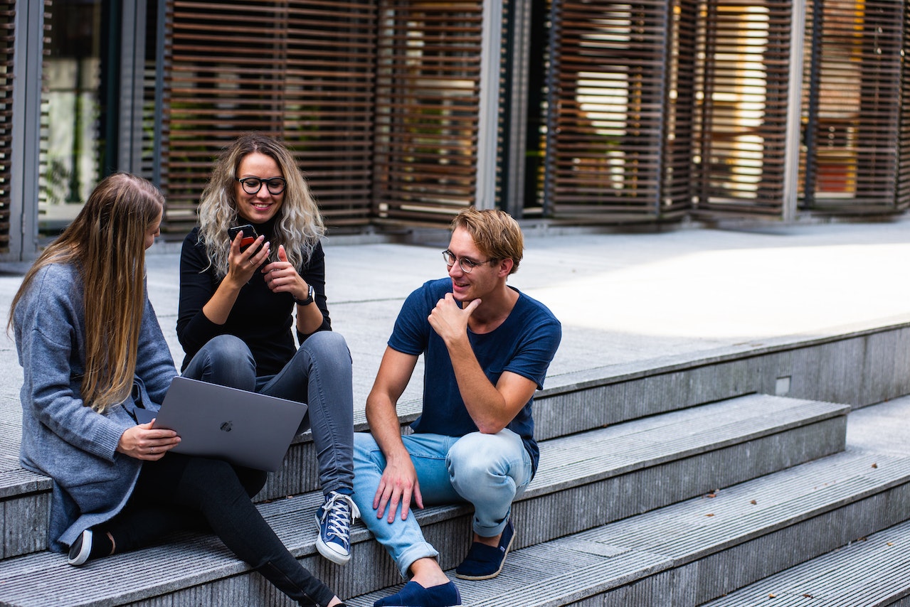 Three college students sitting on starirs outside of a building looking at a computer.