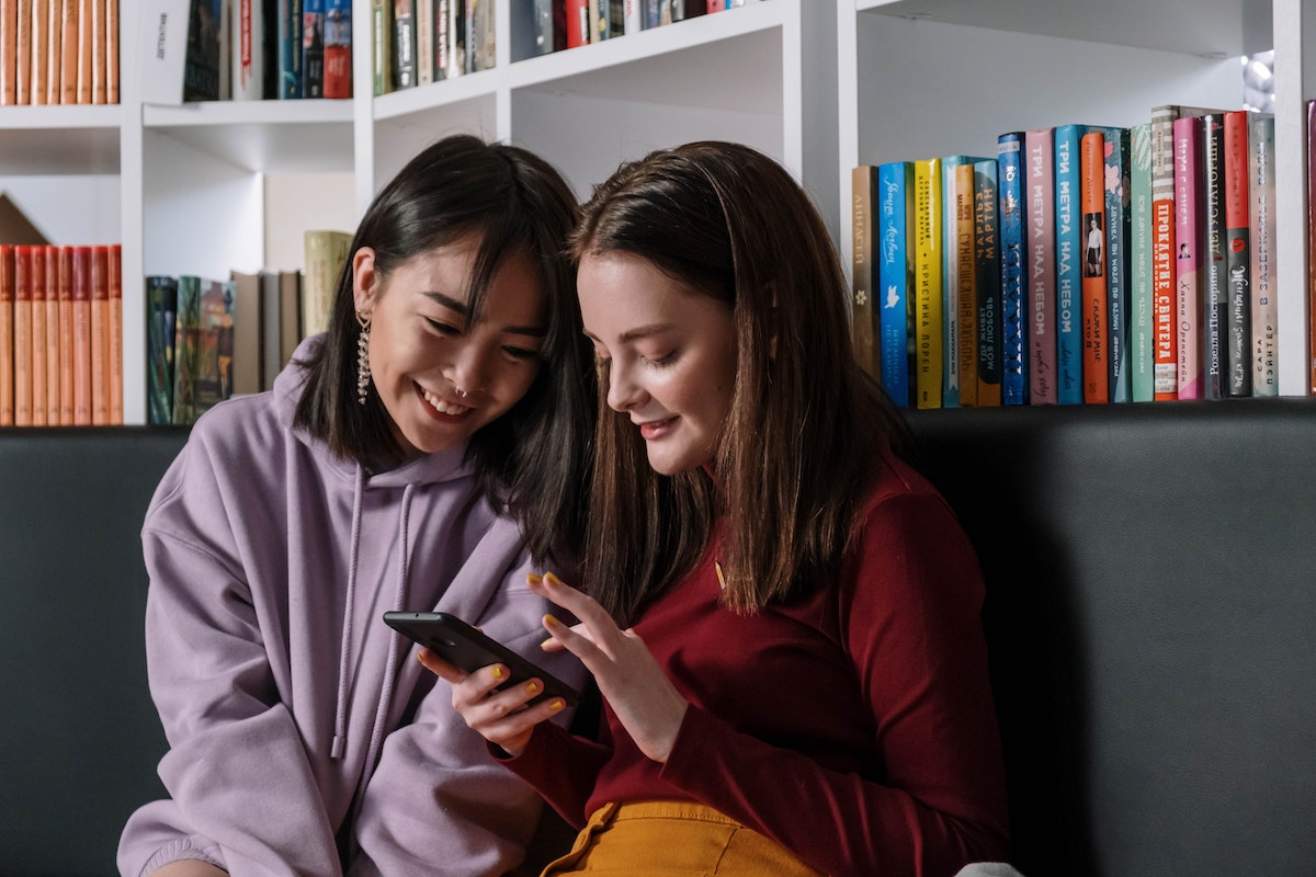 Gen Z girls sitting together in front of bookshelves looking at a phone and smiling.
