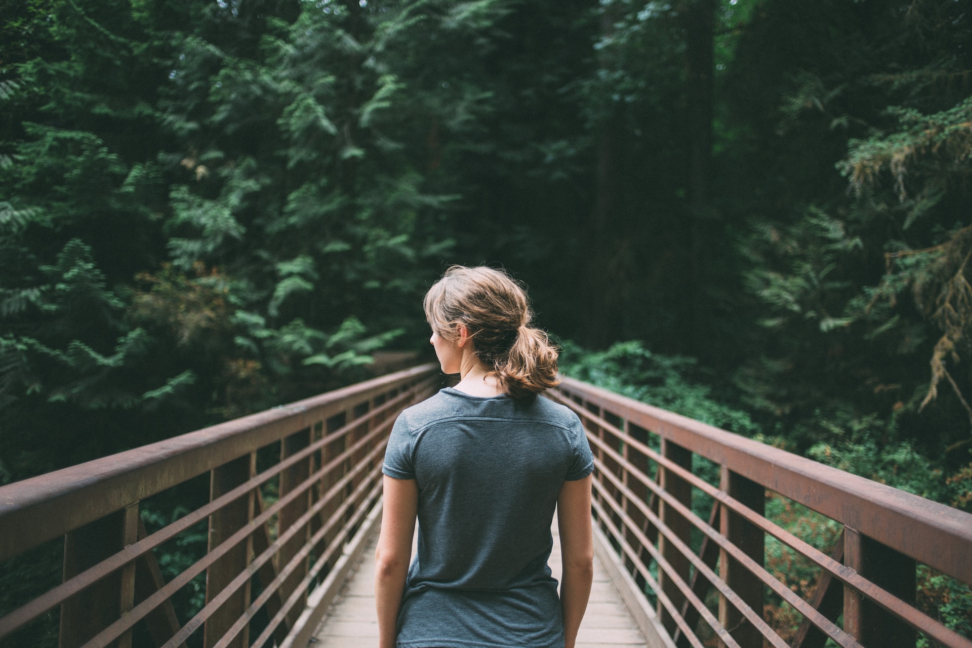 Travel influencer woman with blonde hair and green shirt standing on a wooden bridge in the woods facing away from the camera.