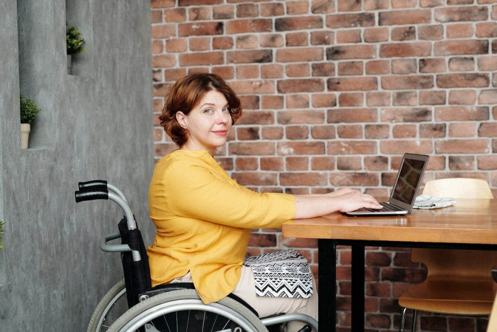 Woman sitting in a wheelchair wearing a yellow shirt and sitting at a desk and working on a laptop computer.