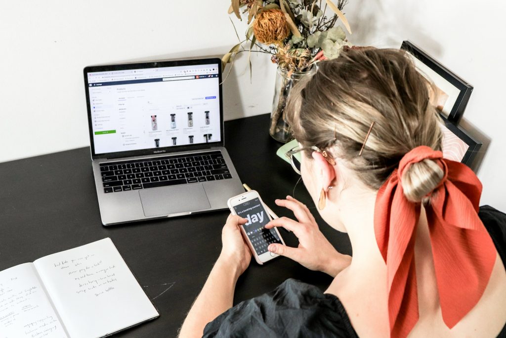 Woman sitting at a desk in front of a laptop and open notebook, editing influencer content on her phone.