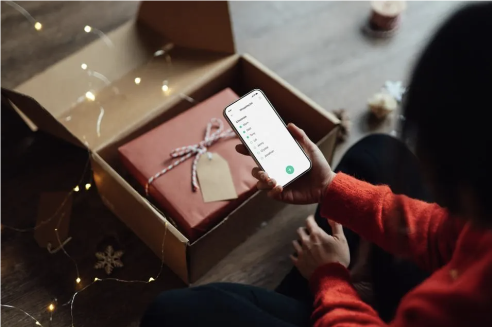 An individual sits on the floor in front of an open gift box and they look at their phone.