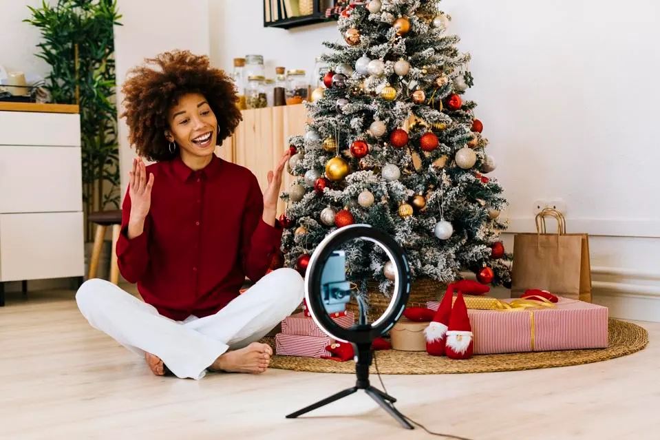 A woman influencer sits cross-legged on her floor in front of a Christmas tree as she speaks to her phone propped up on a ring light.