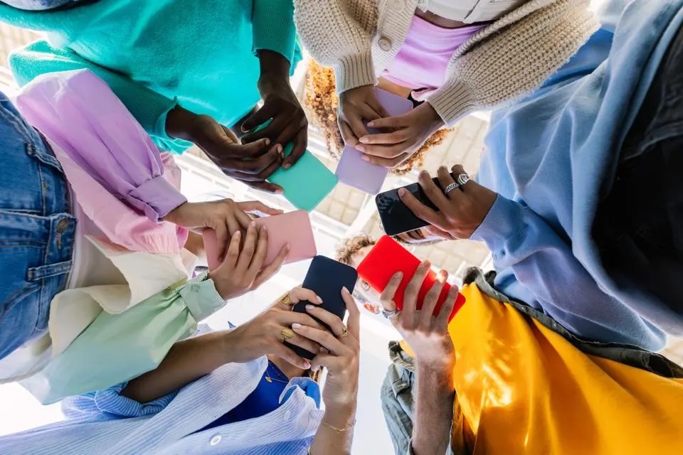 Six people stand in a circle, all looking at their phones. The photo was taken below the subjects, so it appears as though the viewer is looking up at the subjects.