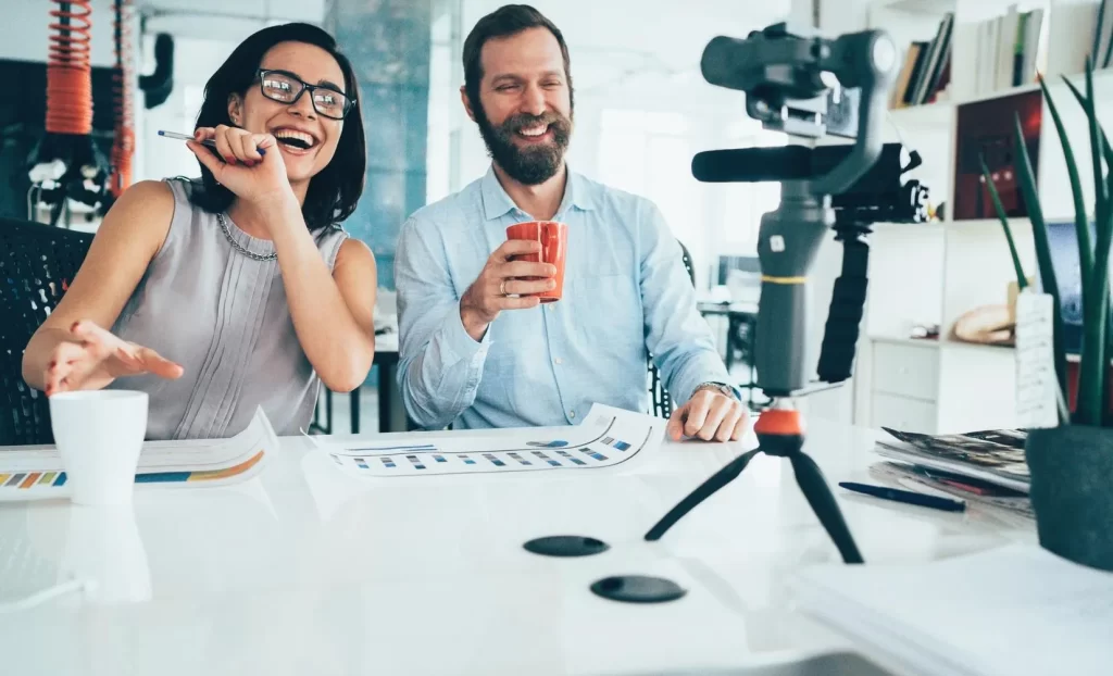 Two people sitting at a desk in front of a camera filming social media content.